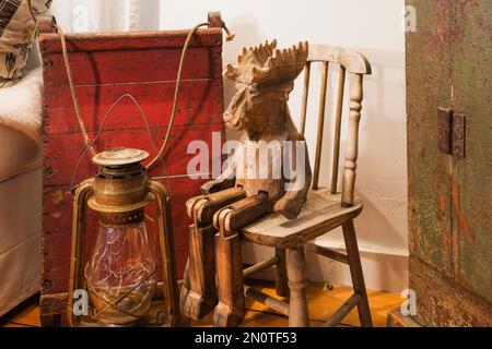 Alte Öllampe, roter Holzschlitten und Elchhölzer Skulptur auf kleinem Stuhl im Wohnzimmer im alten Haus im Cottage-Stil aus den 1862 Jahren. Stockfoto