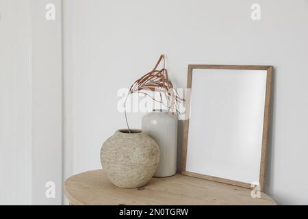 Leeres Holzbilderrahmen-Modell auf altem runden Konsolentisch. Trockenes Palmenblatt in strukturierter Vase. Weißer Wandhintergrund. Leerer Kopierbereich. Elegantes Bo Stockfoto