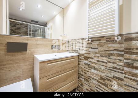 Badezimmer mit weißem Porzellanwaschbecken über der Kommode mit Holzschubladen und rahmenlosem Spiegel an der Wand, passenden Fliesen und weißem beheiztem Handtuchhalter Stockfoto