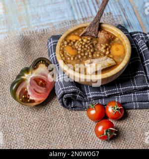 Ein wunderbarer spanischer Eintopf Linsen mit Chorizo in einer Bambus-Holzschüssel auf einem grauen Tuch mit kleinen Tomaten und einer halben reichen Tomate Stockfoto