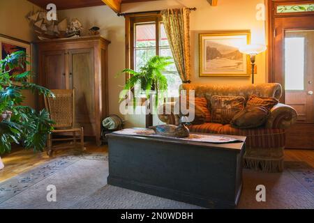 Familienzimmer mit antikem hölzernem Abstellraum Couchtisch, gepolstertem Sofa, gewebtem Sitzschaukelstuhl und Pinienholzschrank in einem alten Haus um 1850. Stockfoto