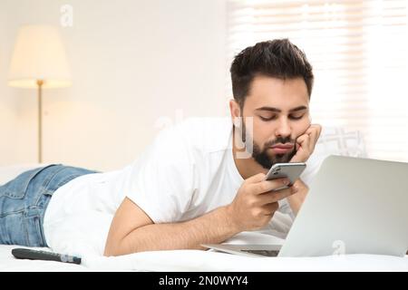 Fauler junger Mann mit Laptop und Smartphone im Bett zu Hause Stockfoto