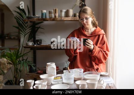 Fokussierte seriöse Geschäftsfrau, Verkäufer von Keramik, Stand in einer Künstlerwerkstatt mit Telefon Stockfoto