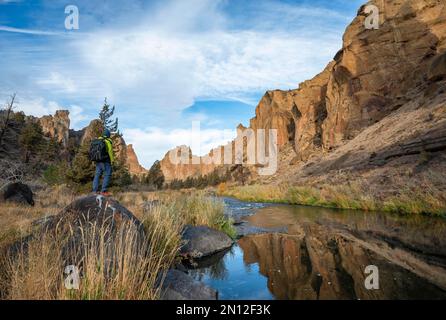 Junger Mann steht auf Stein am Ufer, rote Felswände in der Morgensonne, reflektiert im Fluss, Kurs des Crooked River, Canyon mit Stein für Stockfoto