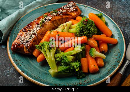 Nahaufnahme von köstlichem gegrilltem Lachsfiletsteak in Teriyaki-Sauce mit geröstetem Gemüse, Karotten und Brokkoli auf einem Teller mit Gabel und Messer auf rustikalem Stil Stockfoto