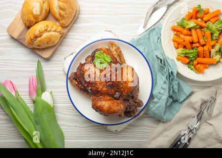 Osteressen auf einem weißen rustikalen Tisch: Gebratenes Hähnchen und Gemüse, Brötchen und Frühlingsblumen Tulpen mit Blick auf Flay Lay, Abendessen mit der Osterfamilie mit fe Stockfoto