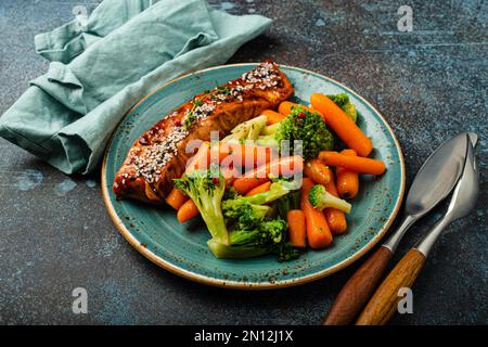 Nahaufnahme von köstlichem gegrilltem Lachsfiletsteak in Teriyaki-Sauce mit geröstetem Gemüse, Karotten und Brokkoli auf einem Teller mit Gabel und Messer auf rustikalem Stil Stockfoto