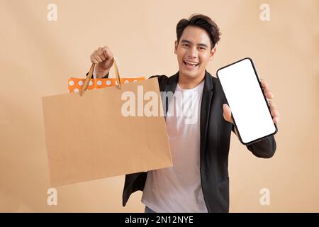 Fröhlicher, lächelnder asiatischer Geschäftsmann mit leerem weißen Smartphone-Bildschirm und vielen Einkaufstaschen auf der anderen Seite modischer fröhlicher Mann in weißem Hemd und Anzug auf beigefarbenem Hintergrund. Stockfoto