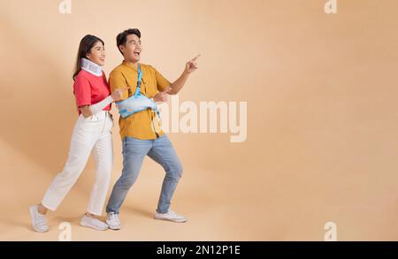 Attraktiver junger asiatischer Mann mit gebrochenem Arm in weicher Schiene und Frau mit Halsschiene und Verband, die auf eine leere Textstelle für Werbung und Werbung zeigt. - Ganzkörperlegnth Stockfoto