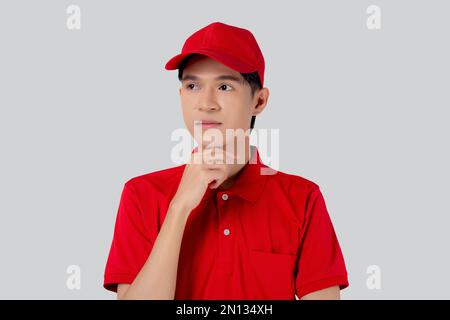 Junger asiatischer Mann in Uniform Rot und Mütze Standing und Denkweise isoliert auf weißem Hintergrund, Mitarbeiter oder Händler, Kurier und Zusteller, Lieferer Stockfoto