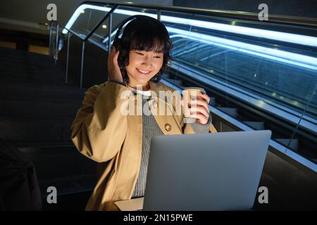 Wunderschöne junge Studentin, koreanische Frau sitzt auf der Treppe, sieht sich Videos auf einem Laptop in Kopfhörern an, trinkt Kaffee zum Mitnehmen, arbeitet an einem öffentlichen Ort Stockfoto