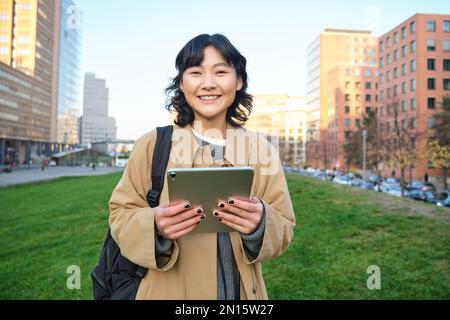 Glückliches asiatisches Mädchen steht auf der Straße, Universitätsstudentin läuft mit einem digitalen Tablet in den Händen und lächelt, steht im Stadtzentrum Stockfoto