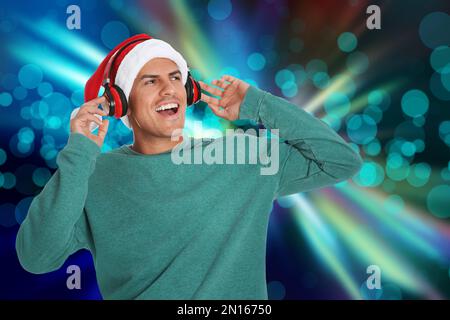 Glücklicher Mann mit Weihnachtsmann-Hut, der Weihnachtsmusik mit Kopfhörern auf hellem Hintergrund und Bokeh-Effekt hört Stockfoto