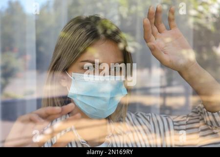 Gestresste Frau mit Schutzmaske, die aus dem Fenster schaut, Blick durch Glas. Selbstisolierung während der COVID-19-Pandemie Stockfoto