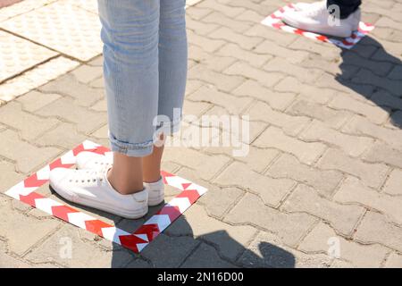 Menschen, die auf festgeklebten Fußbodenmarkierungen für soziale Distanz im Freien stehen, Nahaufnahme. Coronavirus-Pandemie Stockfoto