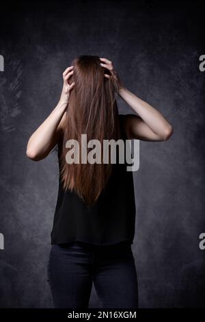 Junge Frau mit langen Haaren, die Hände am Kopf hält. Haare bedecken das Gesicht. Grauer Hintergrund. Kopfschmerz und Migräne Konzept. Stockfoto