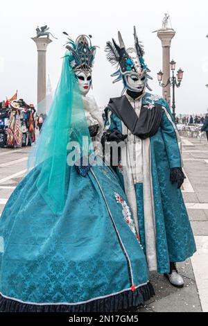 Mann und Frau stehen in roten mittelalterlichen Karnevalskostümen, Hüten und Masken auf der Piazza San Marco in Venedig mit Colonna di San Marco und Colonna di San Todar Stockfoto