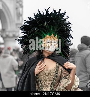 Vinece-Karneval. Eine Frau in einem Vintage-Kostüm mit Spitze, die eine goldene Karnevalsmaske trägt, umgeben von einem Kreis hoher schwarzer Federn, steht allein Stockfoto