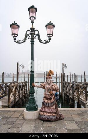 Eine Frau in einem Karnevalskostüm, einer Maske, einem Hut mit Federn, einem Spiegel in der Hand, steht vor dem Hintergrund des Piers auf einem Laternenpfahl Stockfoto