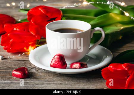 Komposition mit einer Tasse Kaffee, herzförmigen Süßigkeiten und roten Blumen aus der Nähe. Stockfoto