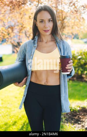 Attraktive Sportlehrerin in Sportbekleidung im grünen Sommerpark mit Rollmatte und einer Plastiktasse Kaffee, die sich nach dem Training ausruhen kann Stockfoto