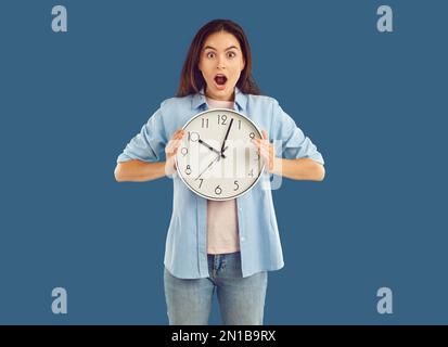 Eine schockierte Frau in lässiger Kleidung hält die Uhr in den Händen auf blauem Hintergrund. Stockfoto