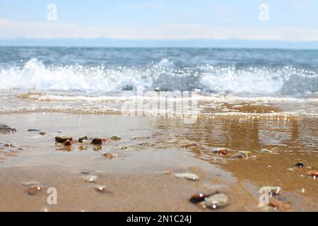 Unschärfe-Strand mit verwischter Welle und nassem Sand mit Kieselsteinen am blauen Himmel. Meer, Meereshintergrund. Outdoor-Urlaub und Reise-Abenteuer-Konzept Stockfoto