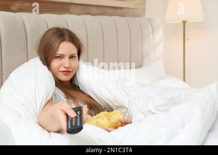 Faule junge Frau mit einer Schüssel Chips, die im Schlafzimmer fernsieht Stockfoto