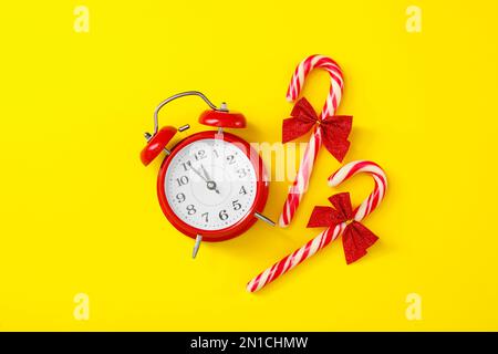 Wecker und Zuckerstangen auf gelbem Hintergrund, flach liegend. Der Countdown für das neue Jahr Stockfoto