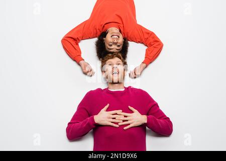Draufsicht auf einen glücklichen rothaarigen Mann in magentafarbenen Kleidern und eine aufgeregte afroamerikanische Frau, die auf weißem, Stock Image liegt Stockfoto