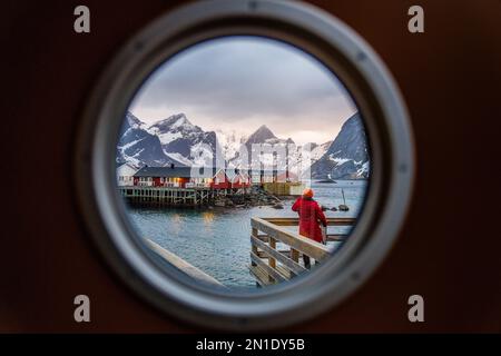 Rückansicht einer Person, die traditionelle Rorbu-Hütten durch ein Bullauge, reine Bucht, Lofoten-Inseln, Norwegen, Skandinavien, Europa Stockfoto