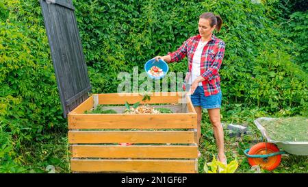 Handgefertigte Kompost-Mülleimer aus Holz – Nahaufnahme. Ein Gärtner in einem karierten Hemd wirft Rasenschnitt und Küchenabfälle in einen Kompost für Heimwerker, um die Fruchtbarkeit zu verbessern Stockfoto