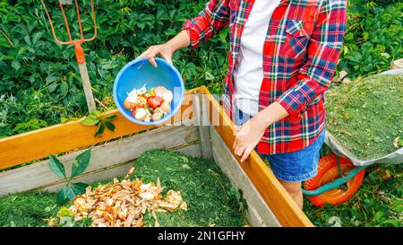 Küchenabfälle, Rasenschnitt als Kompostierungszutaten nähern sich. Eine Frau wirft Lebensmittelabfälle in einen Holzkompost. Erhaltung des ökologischen Landbaus Stockfoto