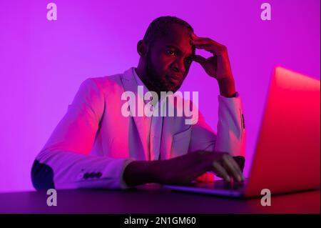 Ein schwarzer Programmierer arbeitet mit einem Laptop. Der Mann arbeitet nachts. Büro mit Neonbeleuchtung Stockfoto