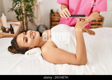 Junge, wunderschöne hispanische Frau, die auf dem Tisch liegt und eine Maniküre im Schönheitssalon bekommt Stockfoto