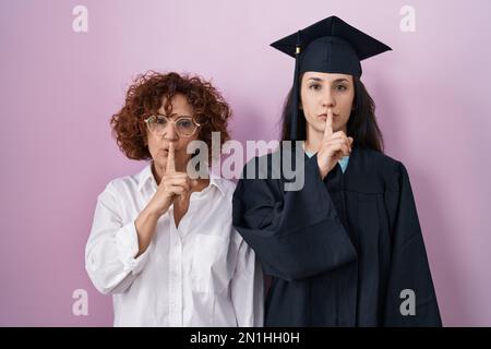 Hispanische Mutter und Tochter tragen Abschlussmütze und Zeremonie-Bademantel und bitten, mit dem Finger auf den Lippen still zu sein. Schweigen und geheimes Konzept. Stockfoto