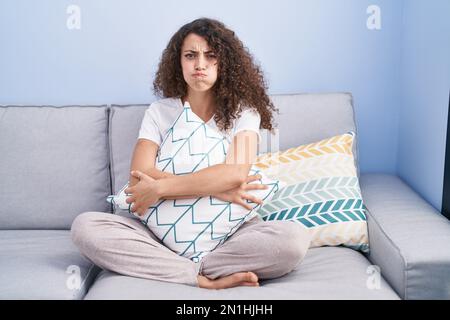Hispanische Frau mit lockigen Haaren, die zu Hause auf dem Sofa sitzt und Wangen mit lustigem Gesicht pustet. Mund mit Luft aufgeblasen, Luft eingefangen. Stockfoto