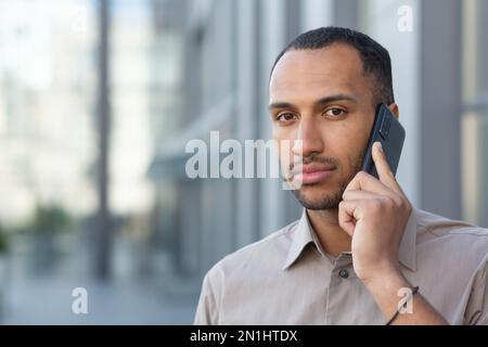 Porträt eines seriösen und fokussierten hispanischen Geschäftsmanns außerhalb des Bürogebäudes, Nahaufnahme eines Mannes, der telefoniert und in die Kamera schaut. Stockfoto