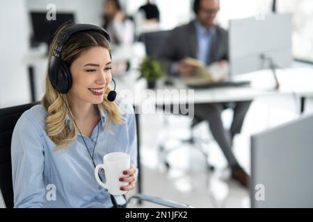 Porträt einer schönen jungen Helpline-Mitarbeiterin, die eine Kaffeetasse hält und sich mit einem Kunden im Callcenter-Büro unterhält Stockfoto