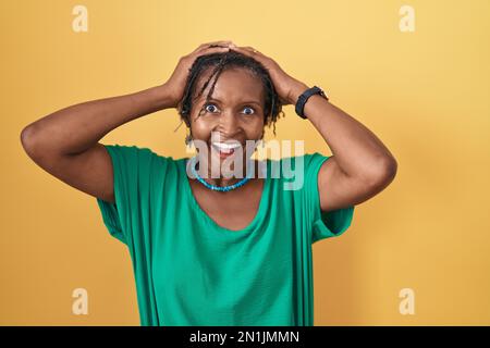 Afrikanische Frau mit Dreadlocks, die auf gelbem Hintergrund steht, verrückt und verängstigt mit Händen am Kopf, ängstlich und überrascht von Schock mit offenem Mund Stockfoto