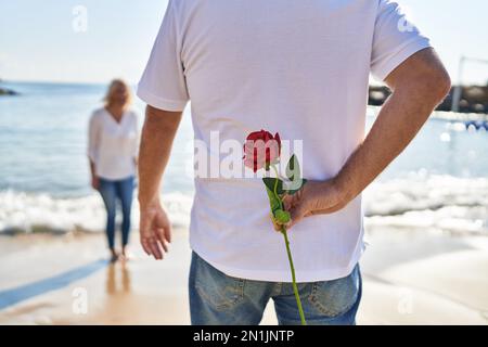 Mann und Frau im mittleren Alter überraschen am Meer mit Blumen auf dem Rücken Stockfoto