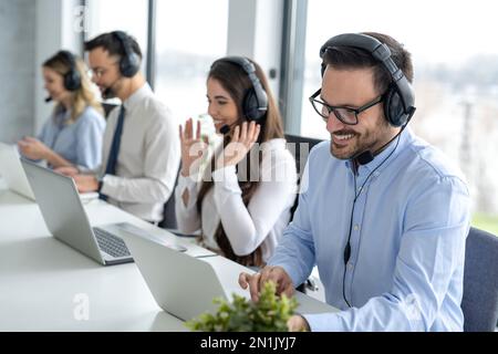 Attraktiver männlicher Mitarbeiter mit Headset, der einen Laptop verwendet und mit anderen Kundendienstmitarbeitern im Büro zusammenarbeitet. Stockfoto
