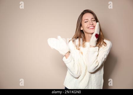 Glückliche junge Frau mit warmem Pullover und gestrickten Fäustlingen auf beigefarbenem Hintergrund Stockfoto