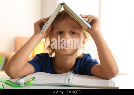 Ein gelangweilter kleiner Junge mit einem Buch auf dem Kopf, der Hausaufgaben macht Stockfoto