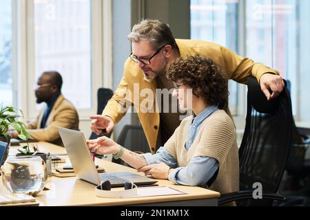 Zwei Büromanager in Freizeitkleidung diskutieren Daten auf dem Laptop-Bildschirm, während eine junge Frau am Schreibtisch sitzt und ein erwachsener Mann sich über sie beugt Stockfoto