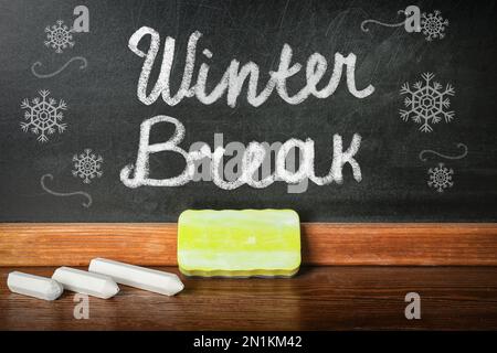 Schreiben Sie Winterferien und Schneeflocken auf die Tafel neben dem Tisch mit Kreide und Staubwedel Stockfoto