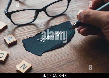 Eine Hand, die My Story schreibt... Auf einem Stück schwarzem Papier. Stockfoto