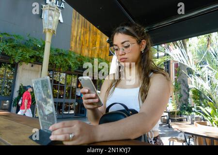 Junge lateinisch-venezolanische Frau mit Brille und langen Haaren, ernsthaft, konzentriert, sitzt in einem Restaurant und scannt den QR-Code mit ihrem Handy Stockfoto