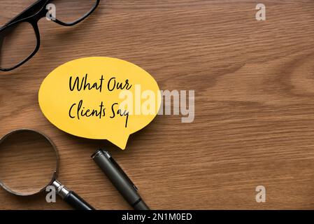 Draufsicht mit Lupe, Brille, Stift und gelber Sprechblase, geschrieben mit „Was unsere Kunden sagen“ auf Holzhintergrund mit Kopierbereich. Stockfoto
