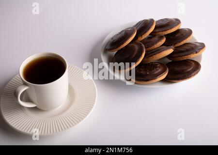 Foto des Kaffees, der in eine weiße Kaffeetasse gegossen wurde und daneben auf einem weißen runden Teller liegt, ist ein mit Schokolade überzogener Keks auf weißem Hintergrund Stockfoto